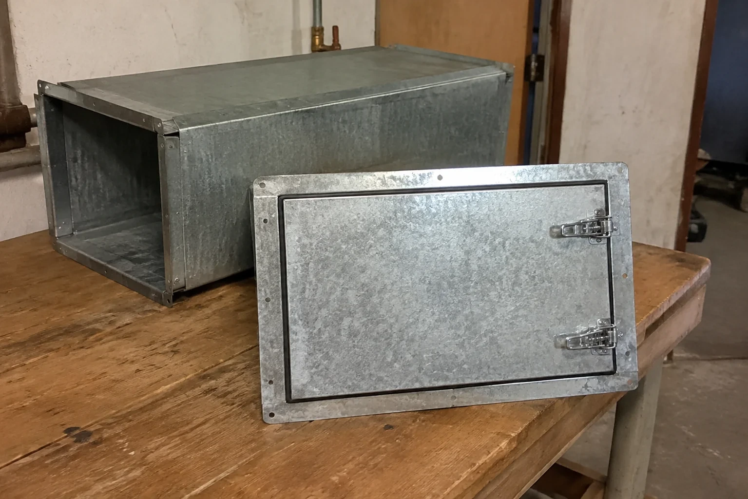 Detached latched metal access panel beside a short rectangular duct section.