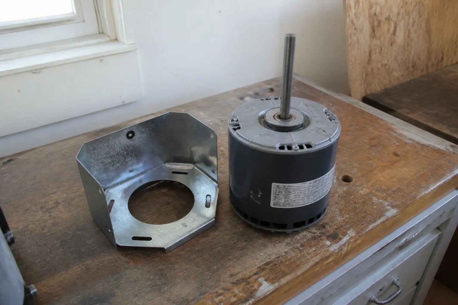 Detached fan motor and metal mounting bracket on a worn workbench.