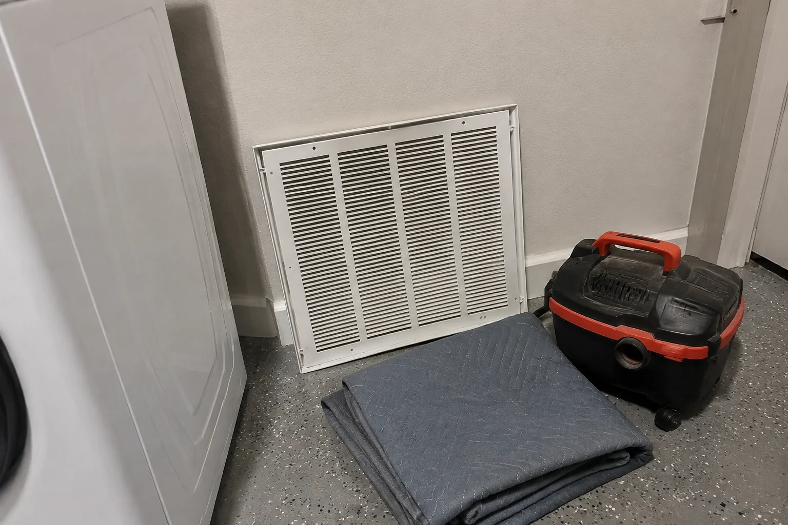 Detached return grille, closed utility vacuum and folded protective cloth in a utility room.