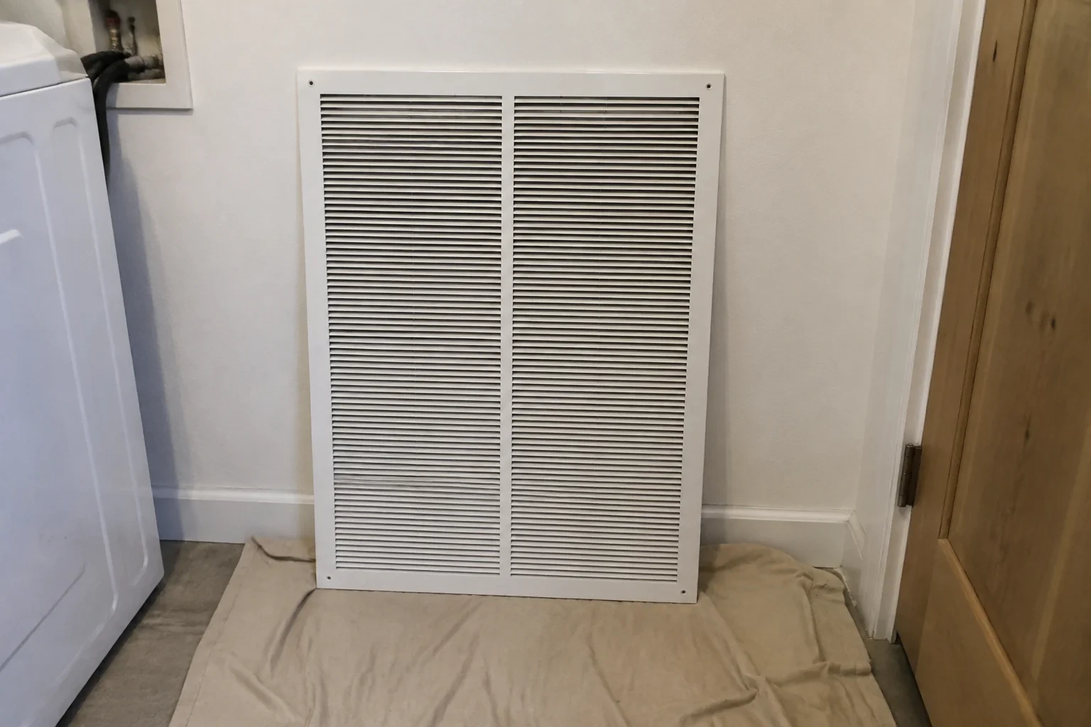 Large detached white return grille resting against a utility-room wall on a protective cloth.